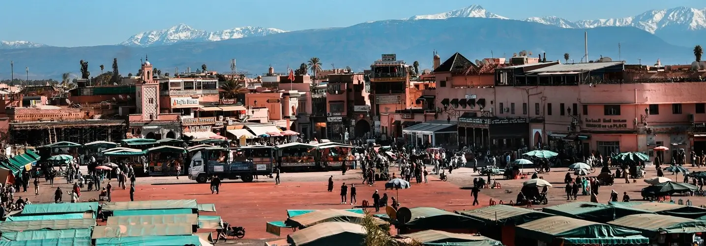 Bustling Jamaa el Fna square with vendors and crowds under umbrellas, set against the backdrop of the snow-capped Atlas Mountains, as part of a 3-day journey from Marrakech to Fes